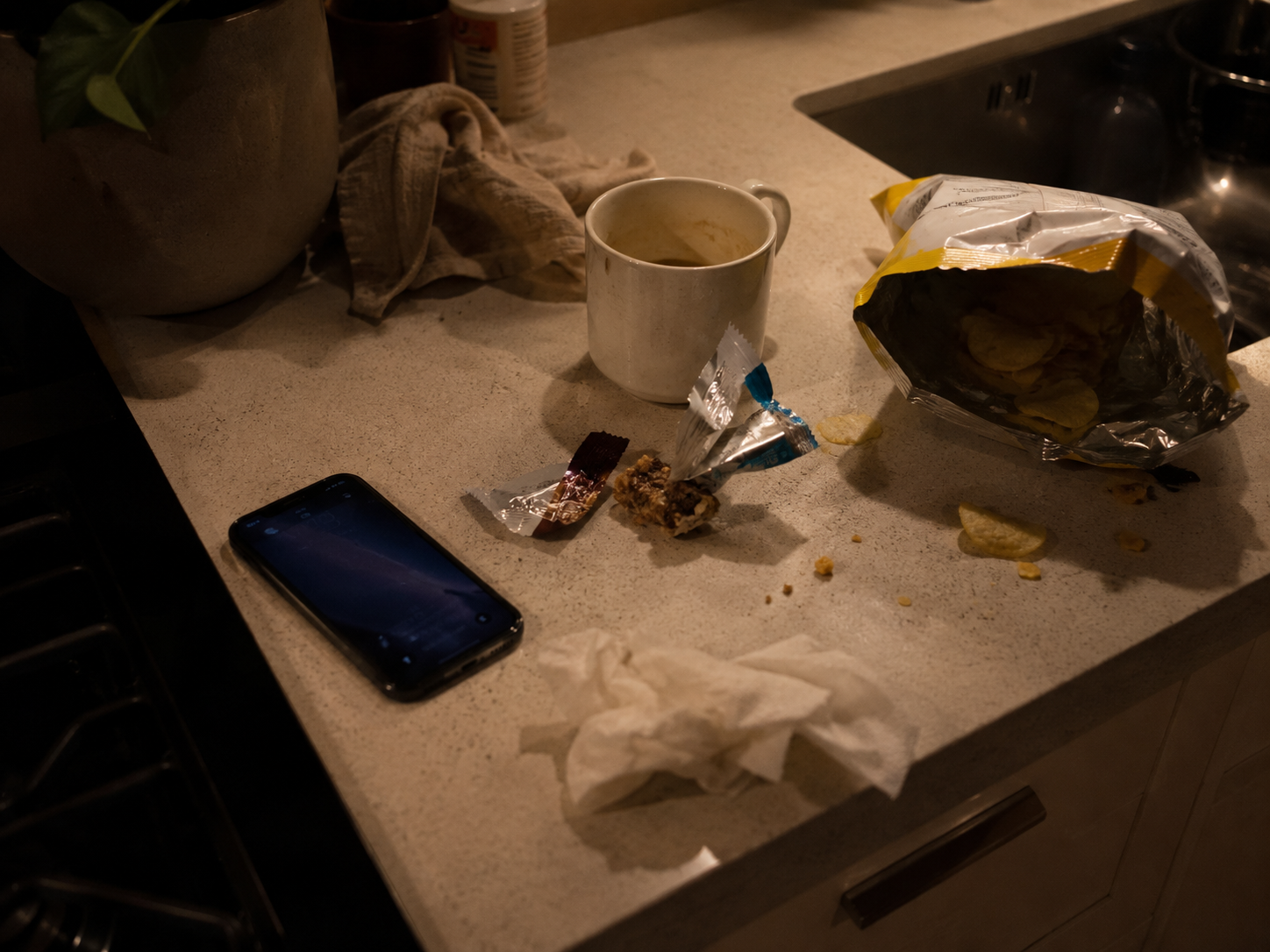 Cluttered evening kitchen counter with a phone and snack wrappers